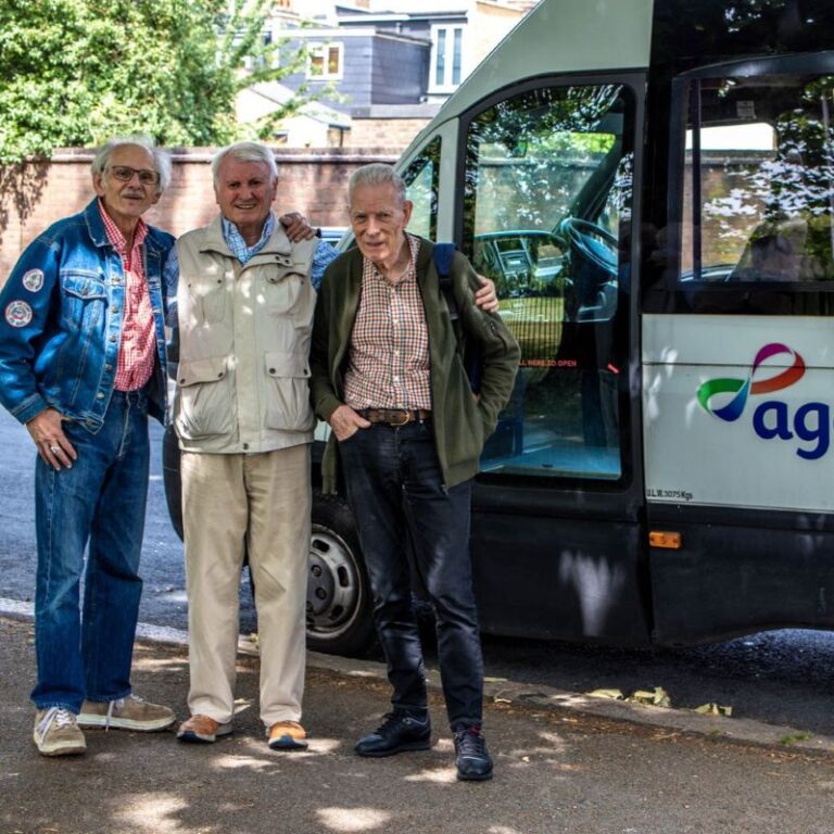 Image shows three older men, age 70+, smiling in front of an AgeUK minibus.