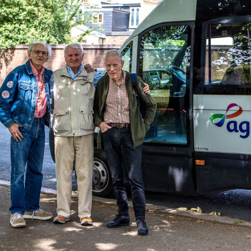 Image shows three older men, age 70+, smiling in front of an AgeUK minibus.