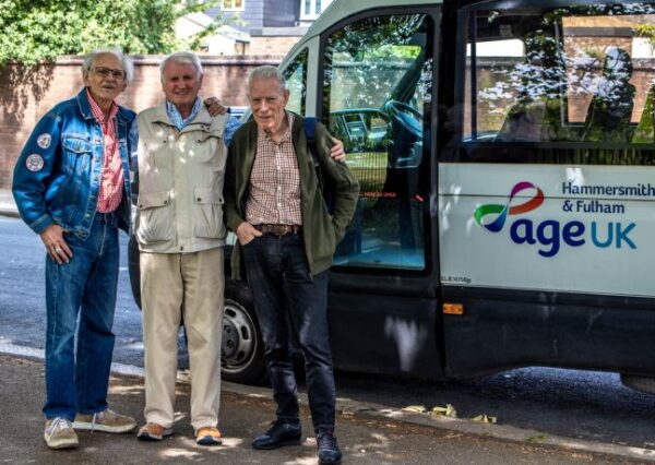 Image shows three older men, age 70+, smiling in front of an AgeUK minibus.