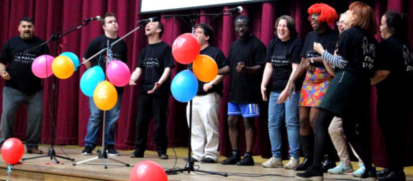 A group of people singing, looking joyful. One woman has brightly coloured red hair. Balloons are on stage with them. 