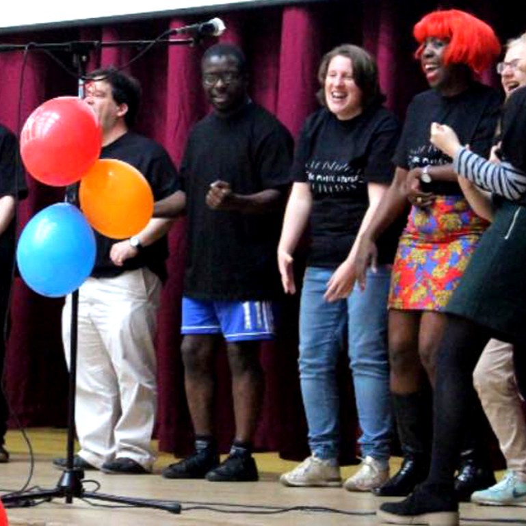 A group of people singing. Balloons are in the foreground. One woman has brightly coloured hair. All look happy.