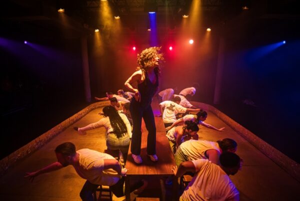 A Bush Theatre performance. One woman stands on a small platform. People around here are kneeling and gesturing to the unseen crowd.