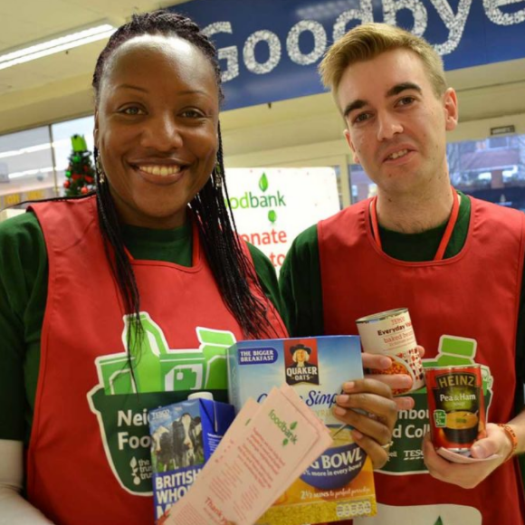 Image shows two people who volunteer for the Hammersmith & Fulham Foodbank. One is a Black woman; the other is a white man. Both are smiling and holding a variety of food that has been donated.