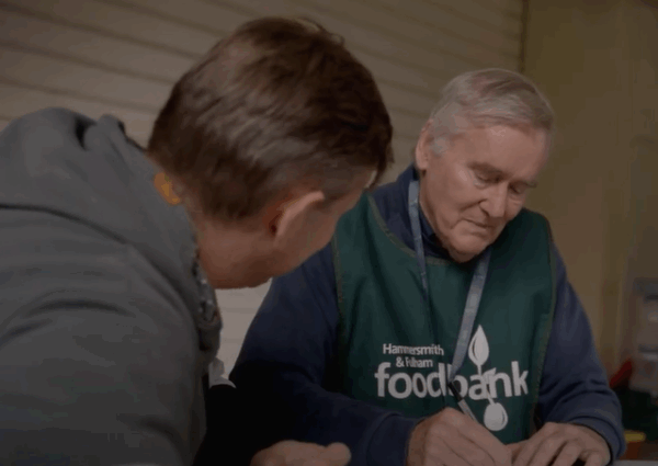 A volunteer for Hammersmith and Fulham Foodbank sits with a client. The volunteer is a white man with grey hair. 