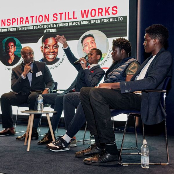 A Black man and three black teenage boys on stage. In the background is a screen saying Inspiration Still Works.