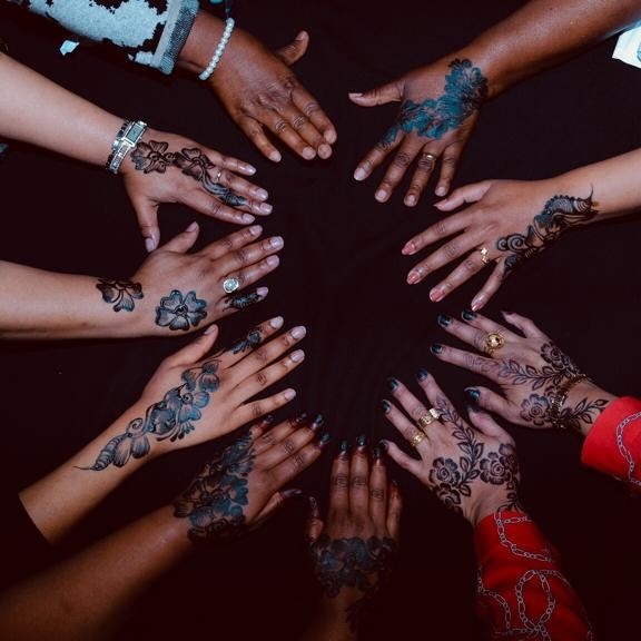 Women's hands facing inwards to create a circle of solidarity. The hands are a range of colours and have beautiful henna patterns on.