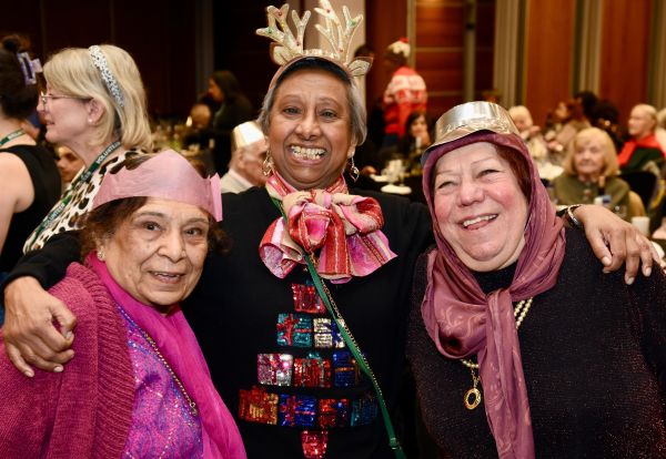 Three women smiling at the camera, One is wearing reindeer horns and a Christmas jumper.  The others are in party hats.