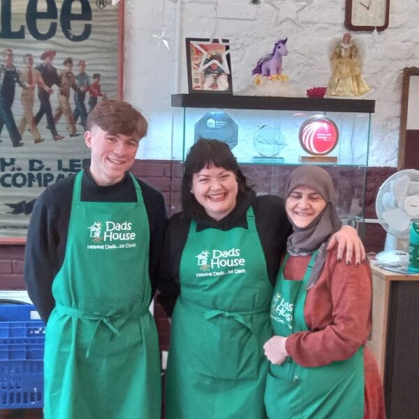 One man and two women wearing Dad's House aprons. They are all smiling and have their arms around each other.