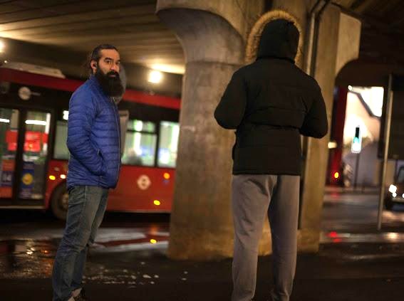 A man and teenage boy talking in the street. The boy has his hood us and back to the camera. 