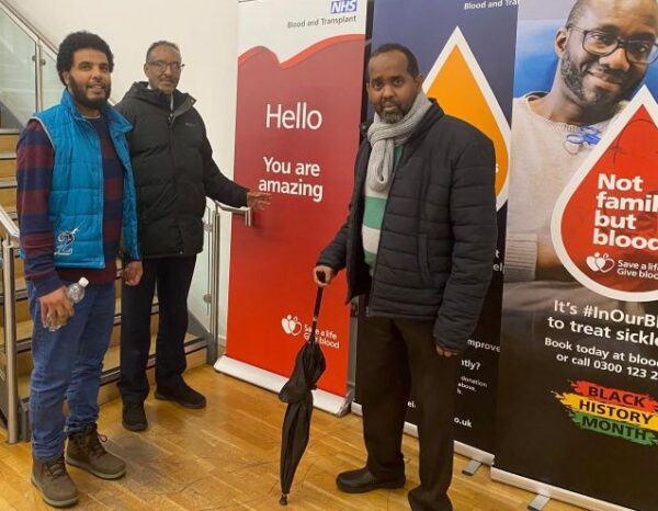 Three men standing in front of a blood donation awareness sign