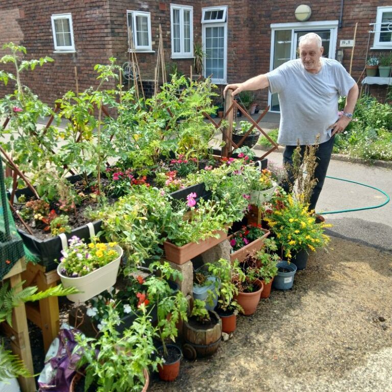 Ken, a man in his 60s, standing by a trug full of plants and surrounded by pots of plants
