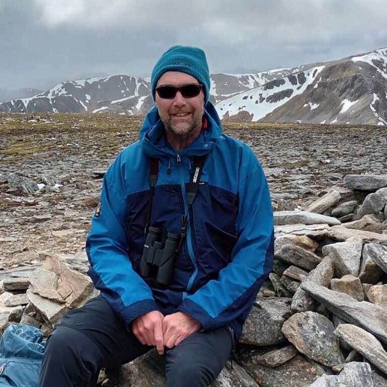 Paul at the top of a mountain wearing a hat, sunglasses and a raincoat.