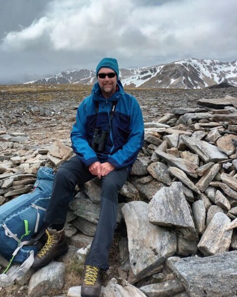 Paul at the top of a mountain wearing a hat, sunglasses and a raincoat. 
