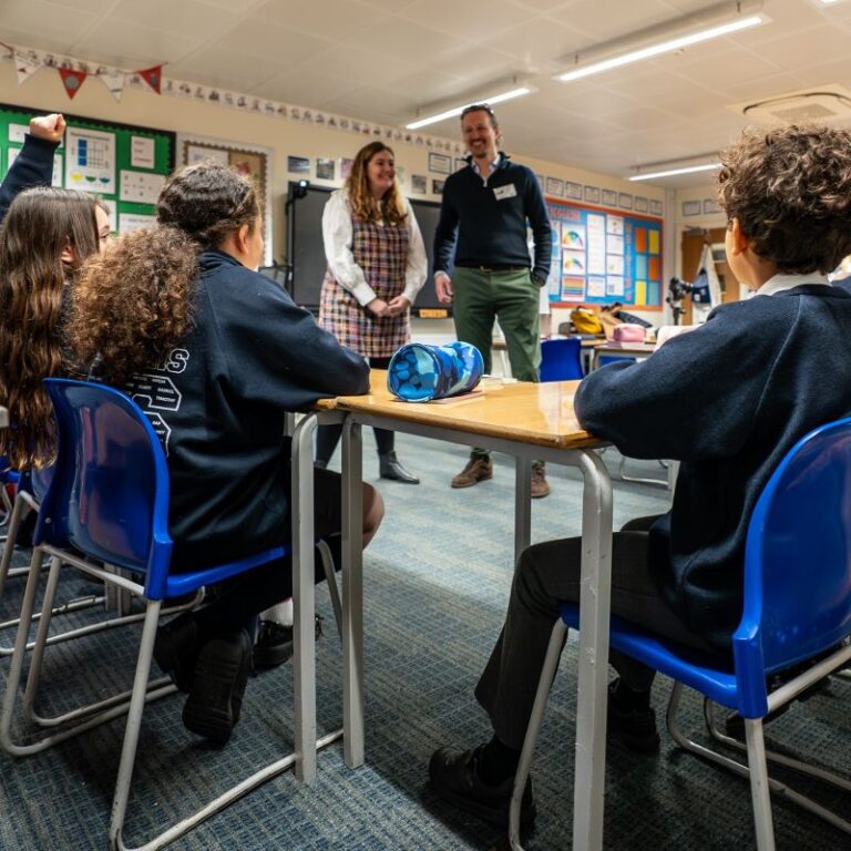 Children in a classroom. They appear to be interested in what the adults are saying. One girl has her hand raised.
