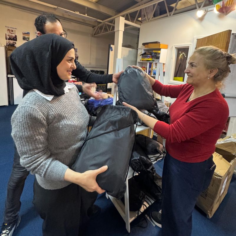 Two women, smiling. One is wearing a headscarf. There is a man standing behind her. They are being handed bags.