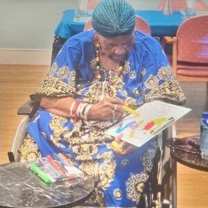 An older woman wearing traditional African dress. She is sat in a wheelchair colouring in a picture.