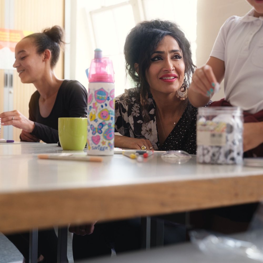 Two women sat at a table. One is looking up at a child standing by her side.