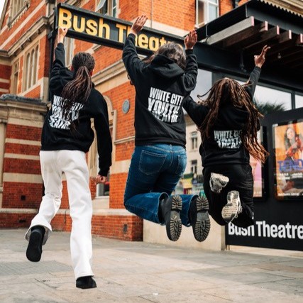 The backs of three girls. They are jumping in the air outside the Bush Theatre. Their hoodies say White City Theatre Project