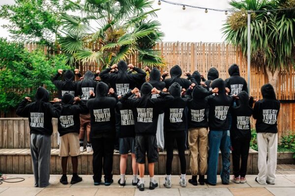 Lots of young people with their backs to the camera. Their hoodies say White City Theatre Project